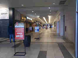 The entrance hallway of the Oak Park Mall. The floor is made of white and gray tile, and the ceiling is lined with potlights and accent lights. Storefronts line the sides, and signs line the middle.