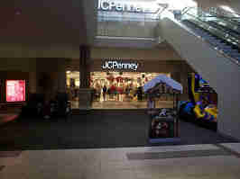 The JCPenney court in the Oak Park Mall from the first floor. The middle of the court has massage chairs, an arcade machine, and a photo prop, with an escalator off to the side.